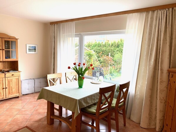 Ein Essbereich mit einem Tisch für vier Personen, Holzsesseln, einer grünen Vase mit Blumen und Blick ins Grüne.