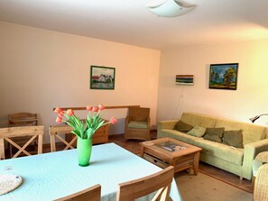 Ein gemütliches Wohnzimmer mit einem Holztisch, einer grünen Vase mit Blumen, einem grünen Sofa und zwei gerahmten Bildern an der Wand.