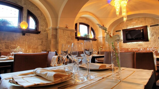 Un restaurante con paredes de piedra, ventanas arqueadas y una araña. Las mesas están dispuestas con copas de vino y platos.