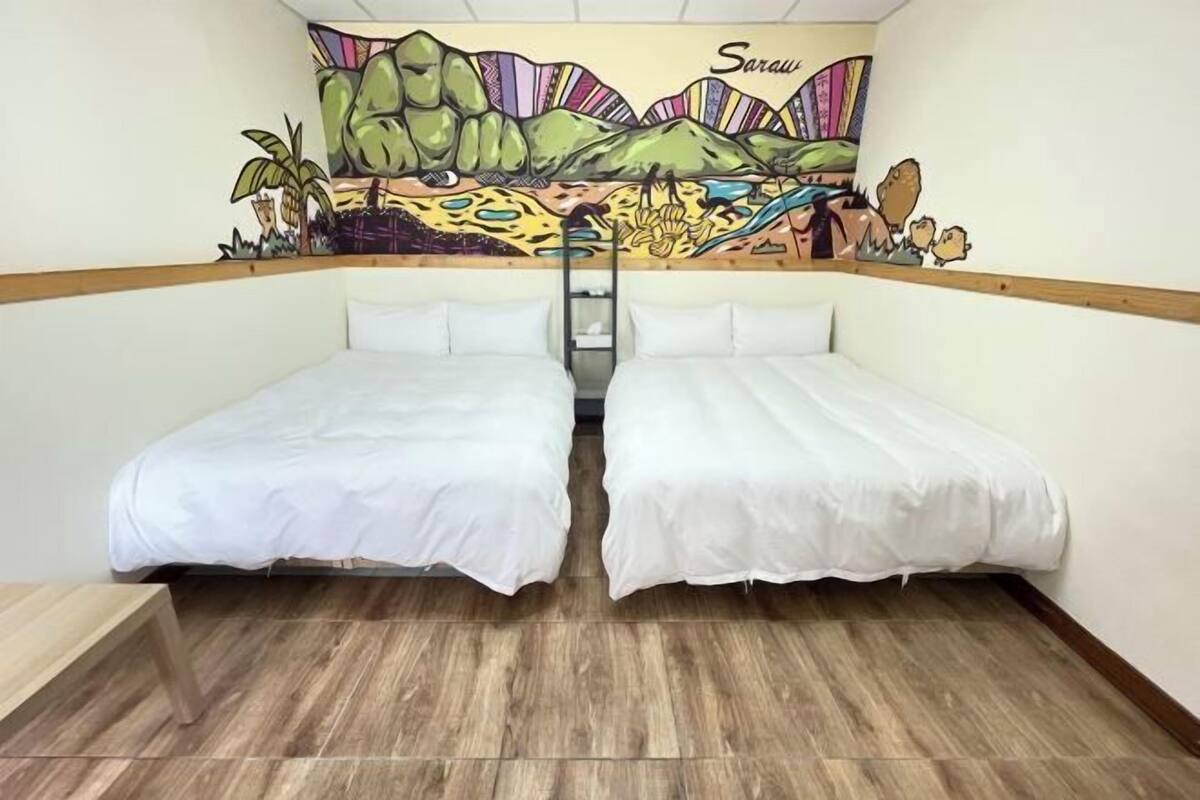 A room with two beds, a wooden floor, and a mural on the wall depicting a landscape with mountains and palm trees.