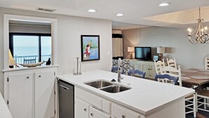 A modern kitchen with a built-in dishwasher, a large island with a sink, and a dining area with a TV and chandelier. - Oceanfront Condo on 7-Mile Beach, Elevator, Gated Peninsula Locale, Pools, Dock (Pawleys Island)
