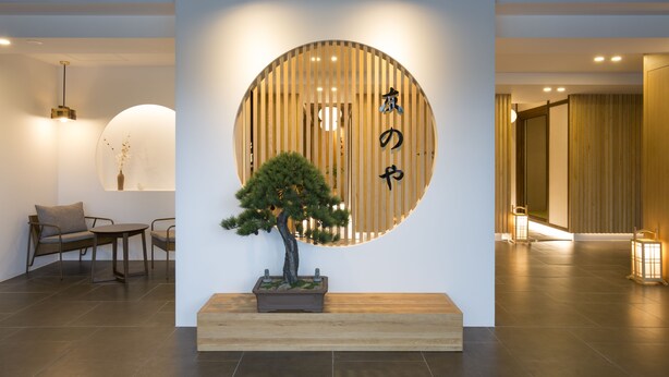 A reception area with a wooden reception desk, a potted bonsai tree, and a circular wooden panel with Japanese characters. - Tomonoya Ryokan (Geoje)