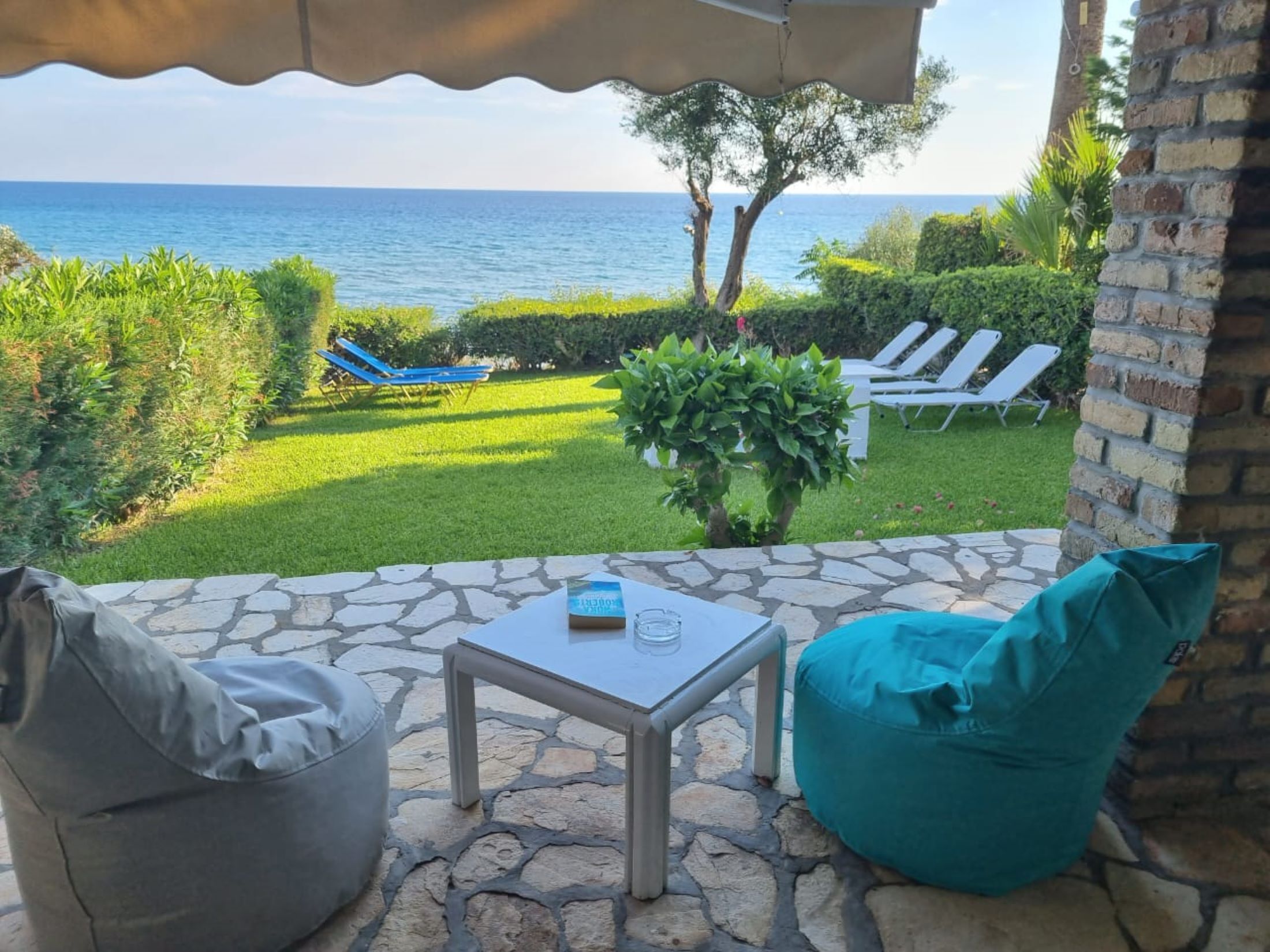 A patio with a stone floor, a table, a teal bean bag, and a view of the sea with lounge chairs.