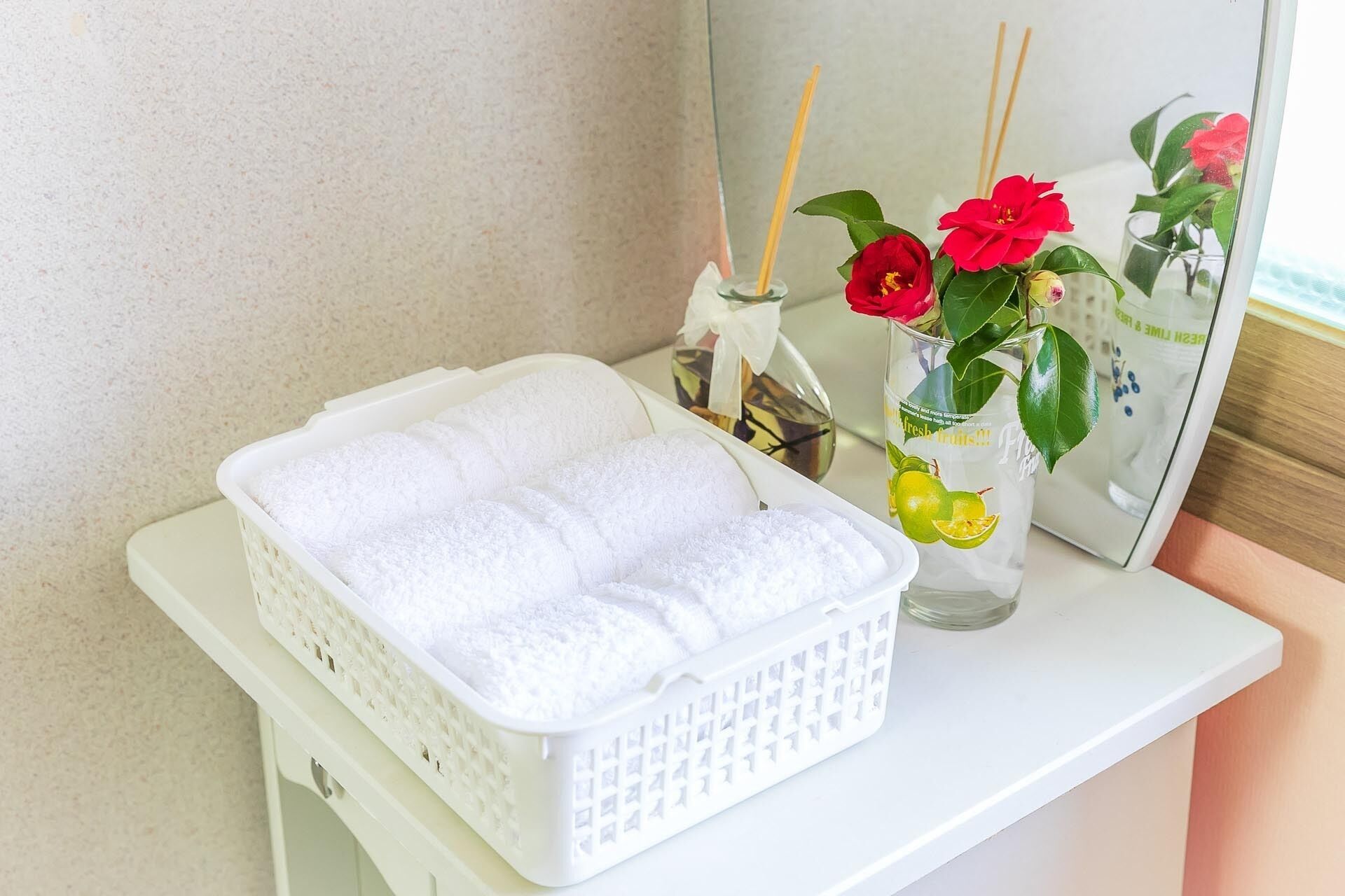 A bathroom with a white sink, a white basket with white towels, a glass vase with red flowers, and a mirror reflecting the flowers and a decorative item.