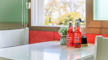 A table with two bottles of red beverage, a small vase with white flowers, and a black appliance on a white table near a window with a view of trees.