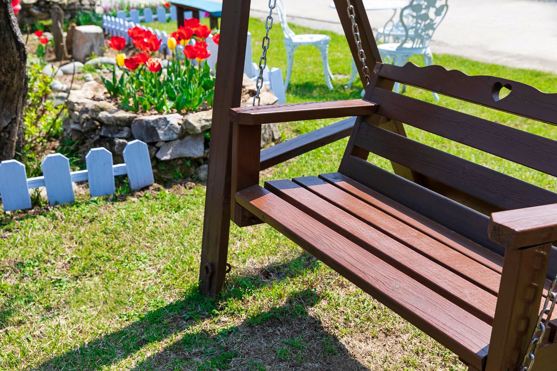 A wooden bench with a metal chain, set in a garden with a white picket fence and red flowers.