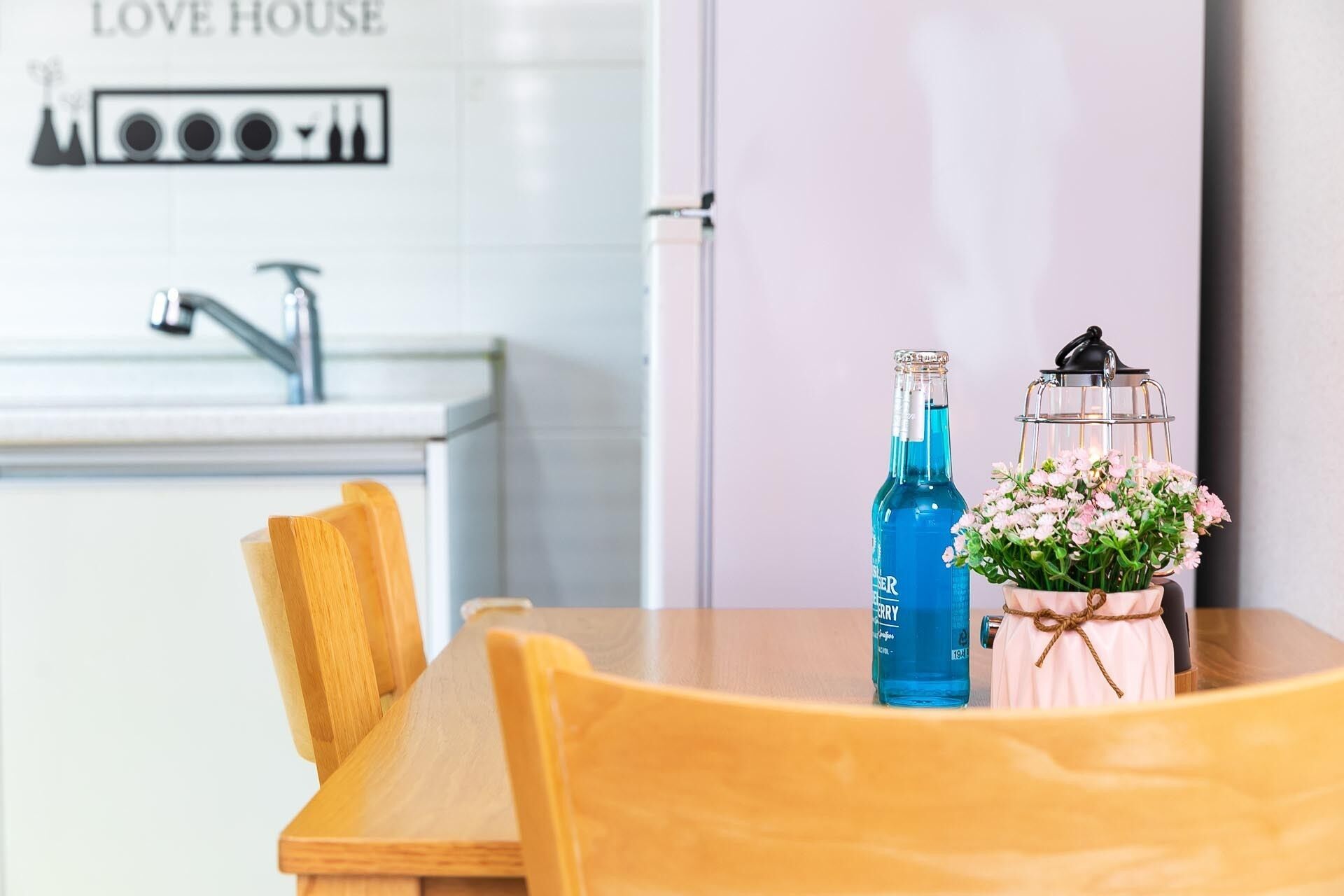 A wooden table with a blue bottle and a lantern with flowers. A kitchen with a sink and a sign reading "LOVE HOUSE" is in the background.