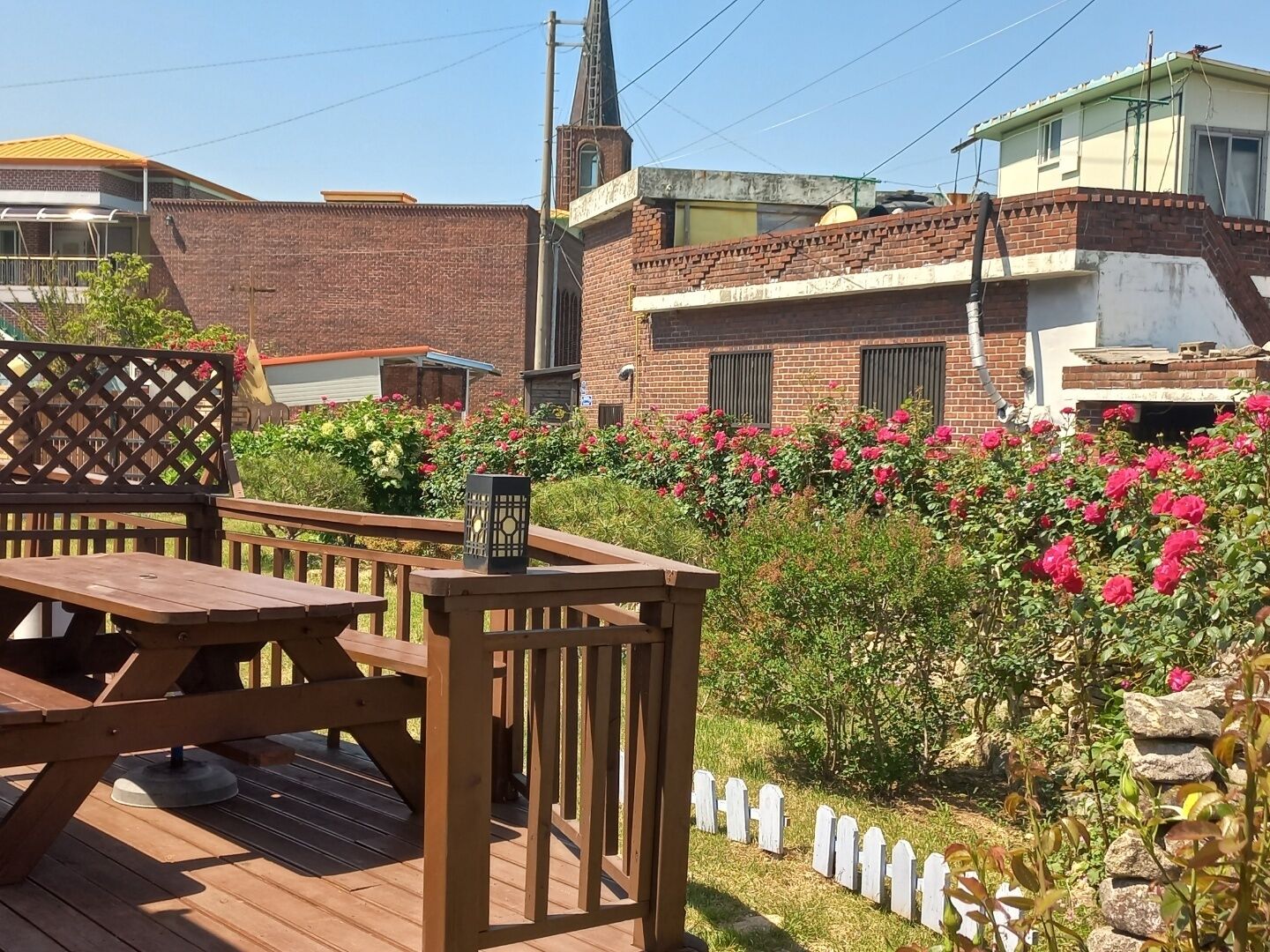A wooden deck with a table and chairs, surrounded by a garden with pink flowers and a white picket fence.