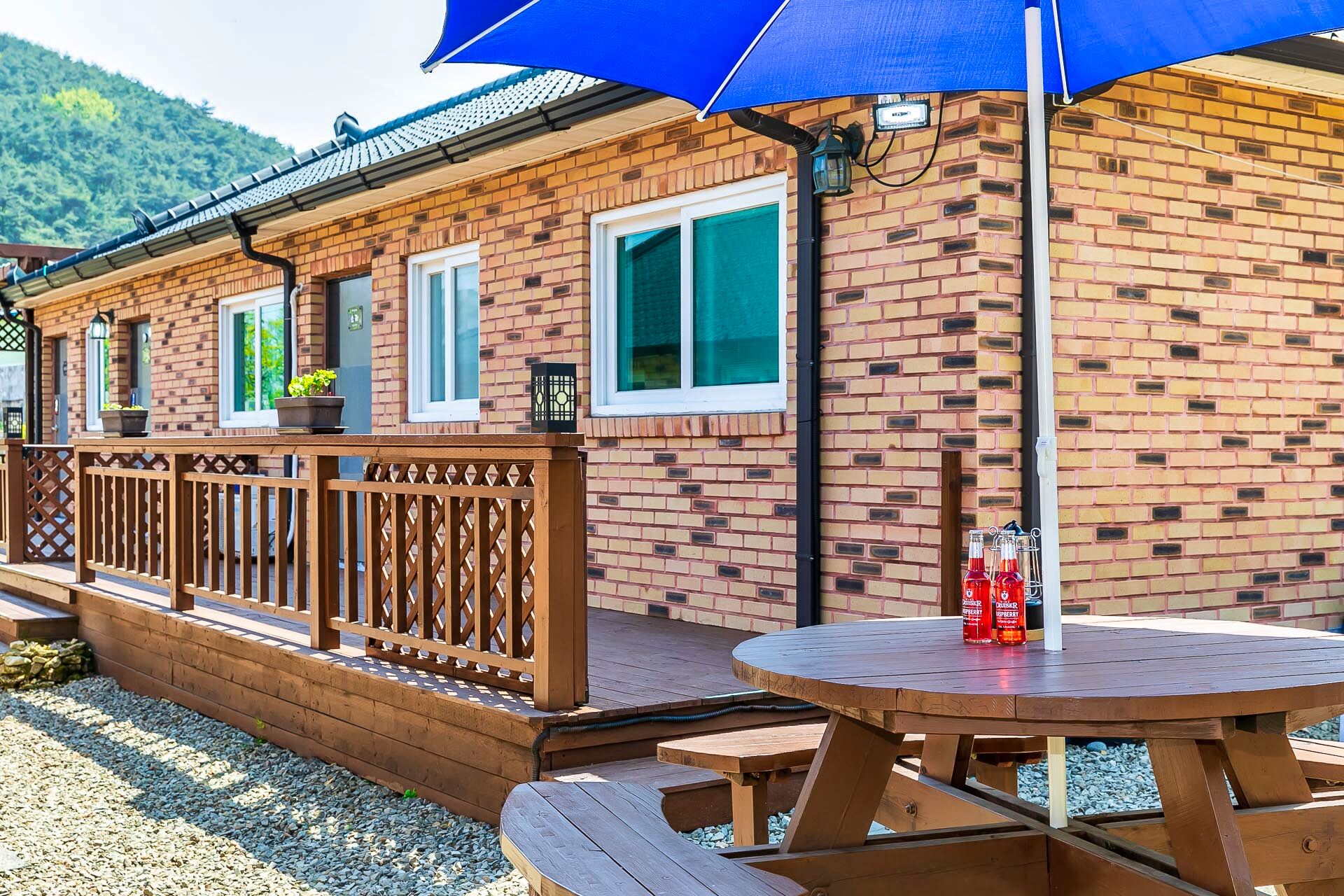 A brick building with a wooden deck, a round table with benches, and a blue umbrella.