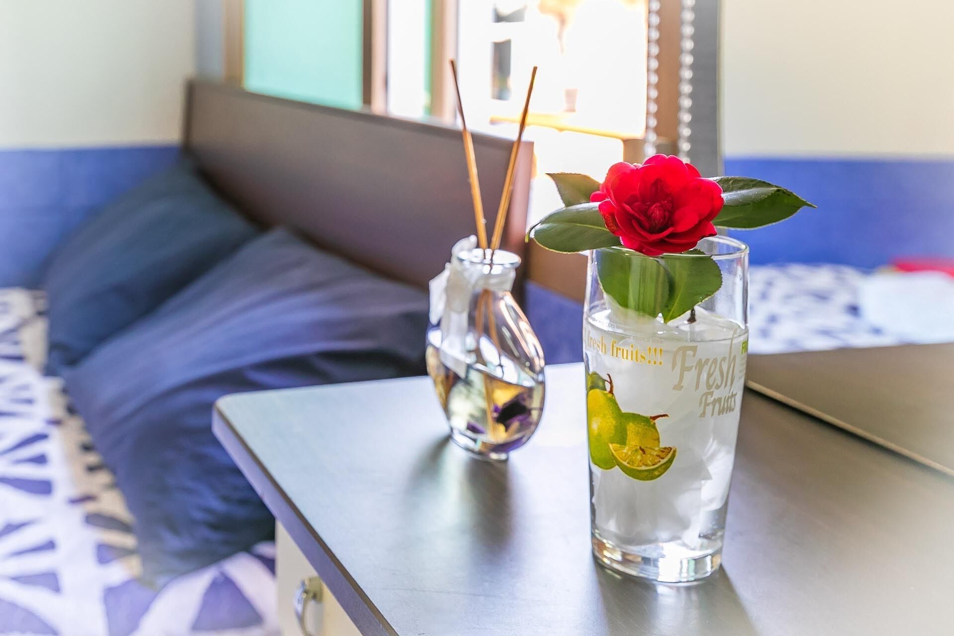 Una habitación con una cama, una mesa, un jarrón con una rosa y un vaso con fruta y agua.