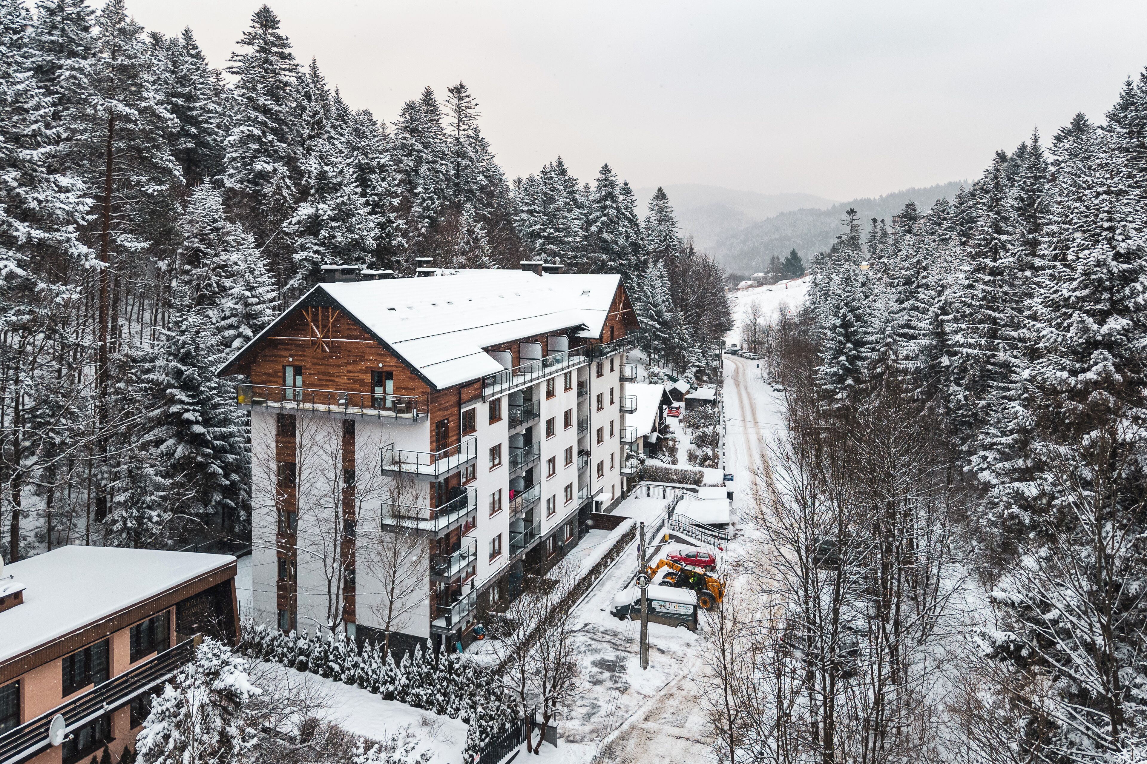 A multi-story building with a wooden facade and a snow-covered roof, surrounded by snow-laden trees and a mountainous backdrop.