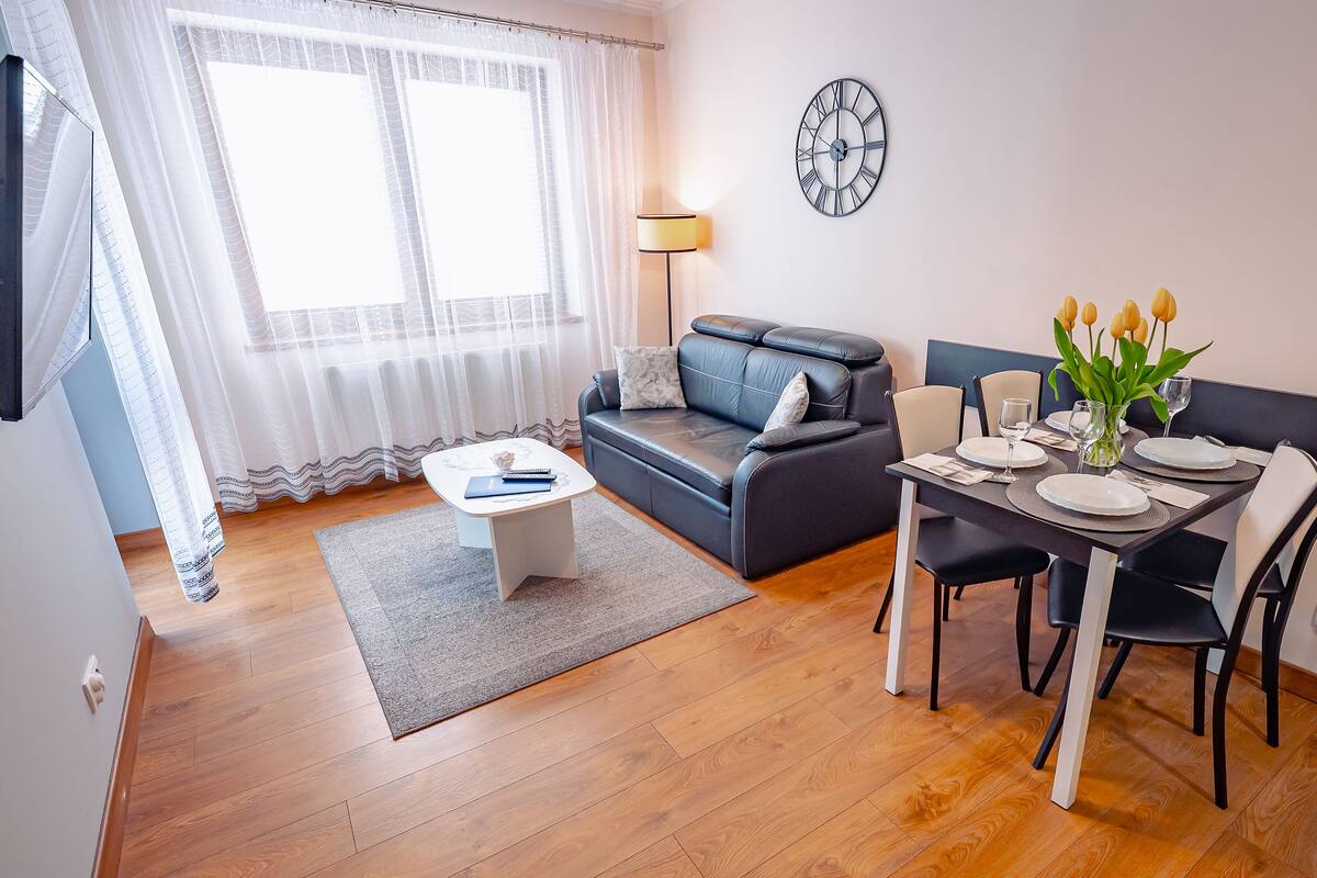 Un salon moderne avec un canapé, une table à manger dressée pour deux personnes et une grande fenêtre ornée de rideaux légers.
