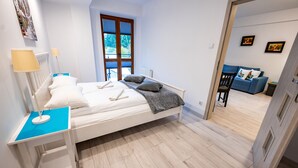 A bedroom with a bed, bedside table, lamp, and a view of a living area with a sofa and a chair.