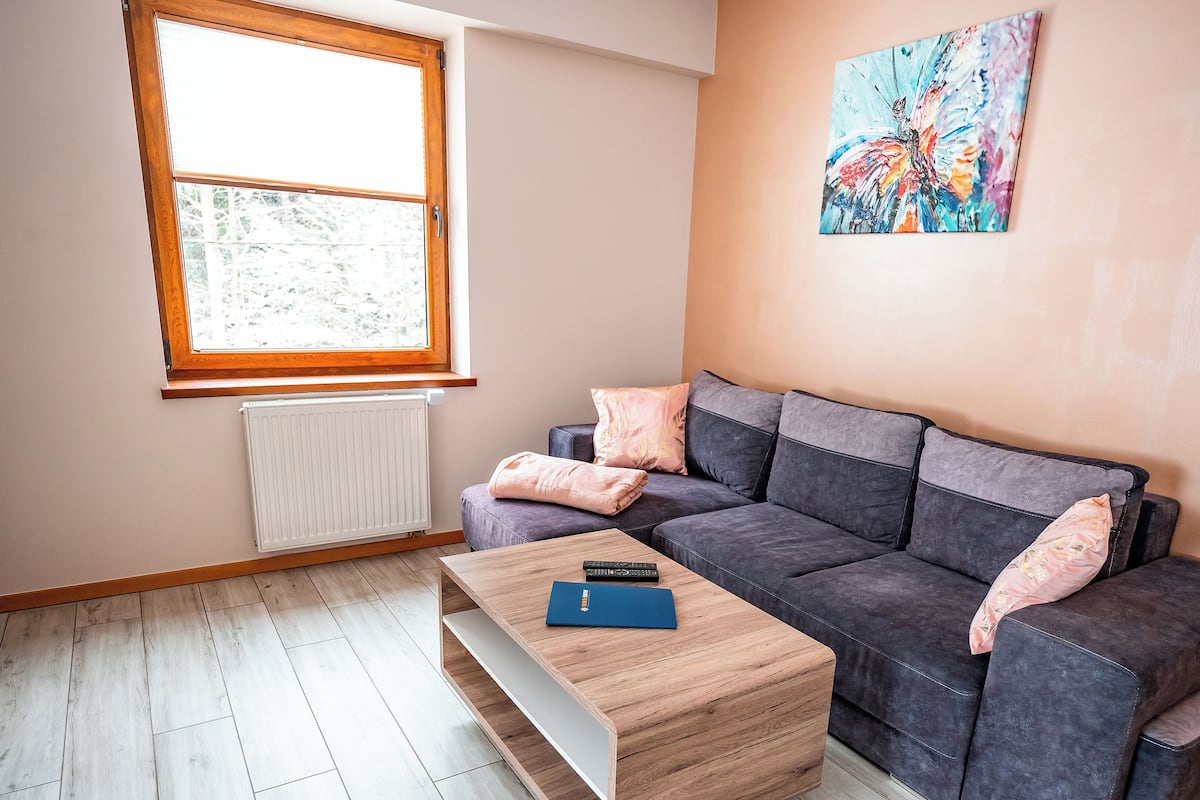 Un salon avec un canapé gris, une table basse en bois, une fenêtre donnant sur des arbres et un tableau abstrait aux couleurs vives accroché au mur.