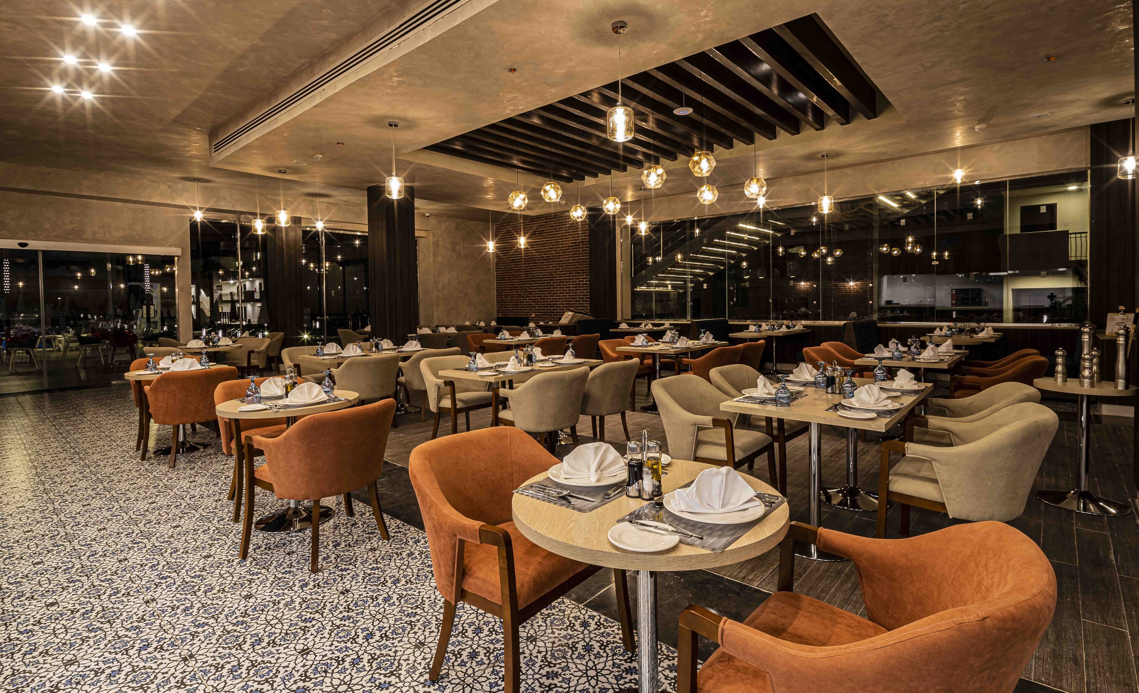 Een restaurant met ronde tafels en oranje stoelen, met een patroon vloer en hangende verlichting.