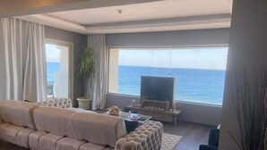 A living room with a sofa, a TV, a plant, and a view of the sea.
