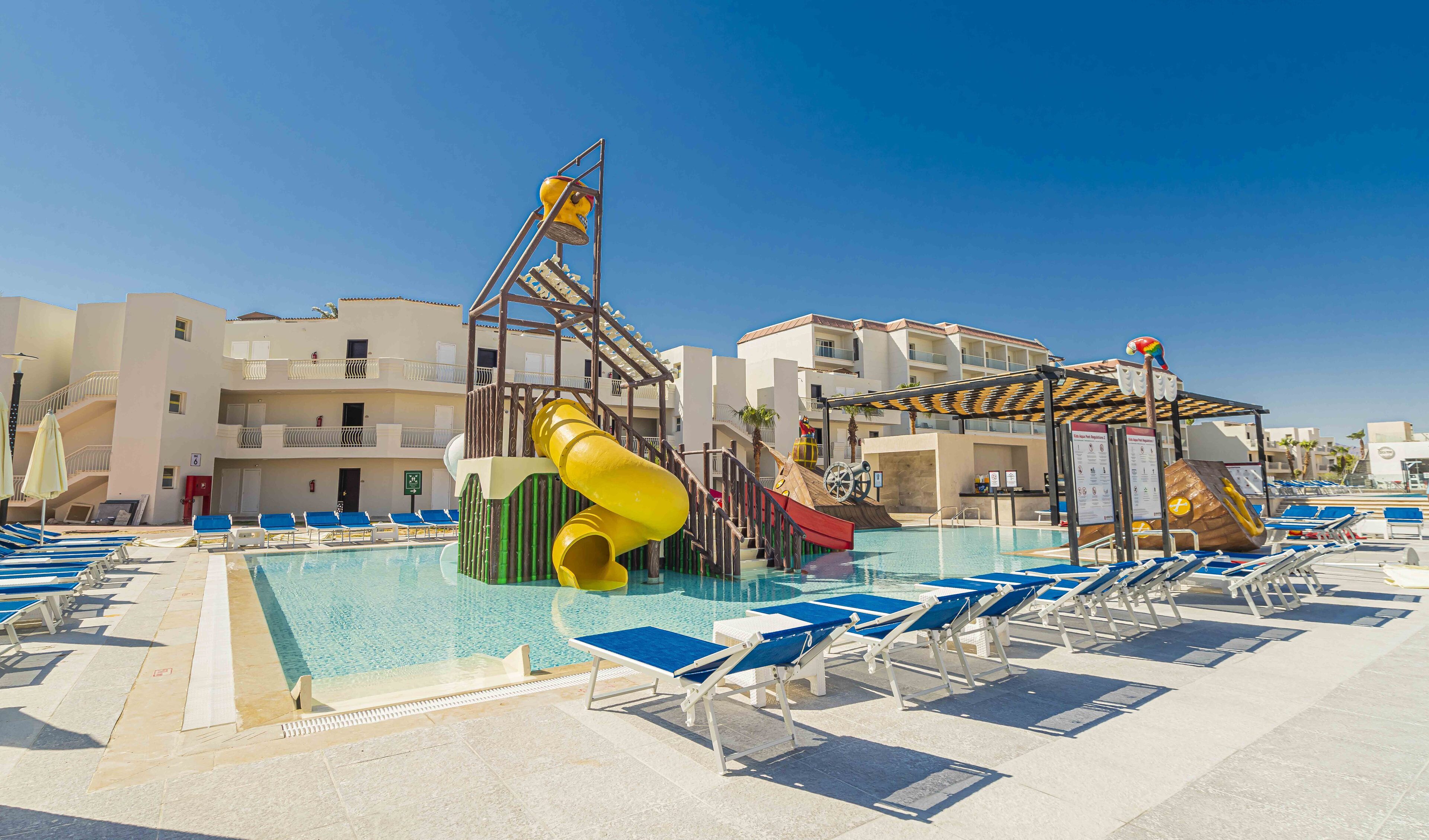 A pool area with blue lounge chairs, a yellow slide, and a water play structure.
