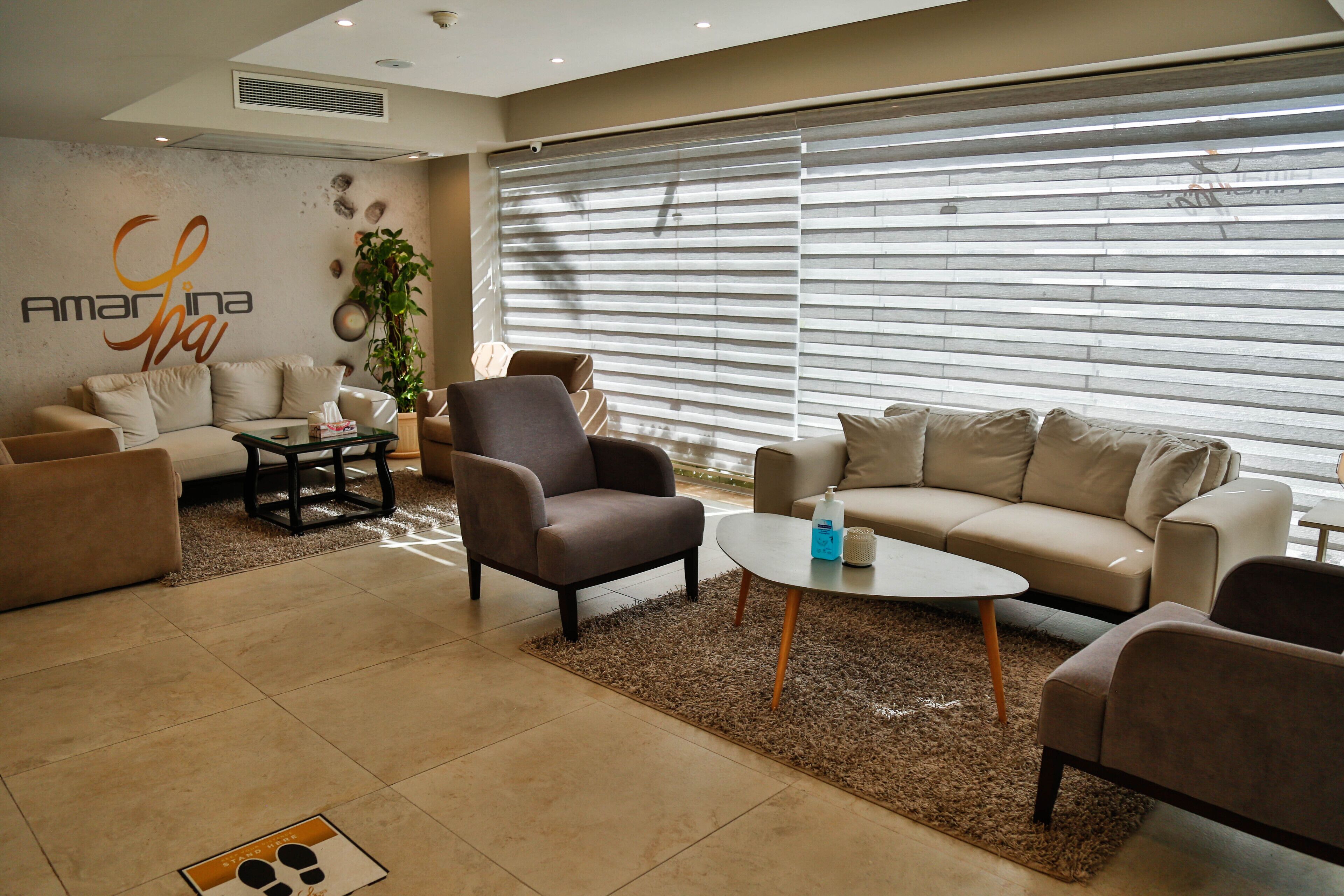 Un hall d’hôtel moderne avec un canapé beige, des fauteuils, une table basse et une grande fenêtre équipée de stores.