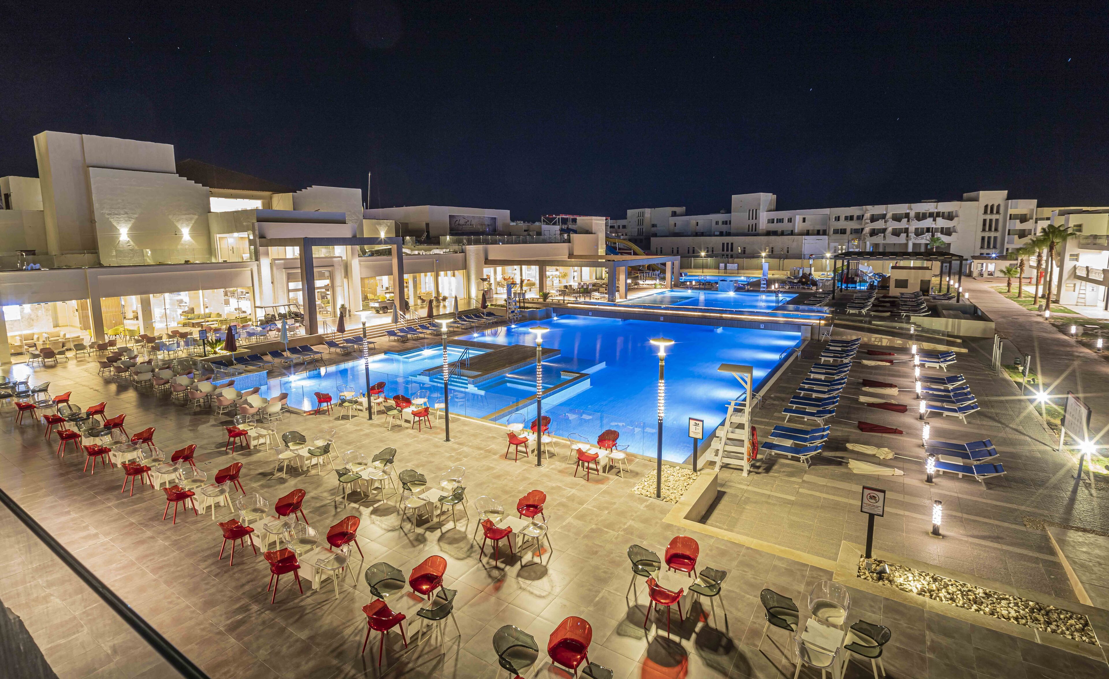 A hotel pool area with multiple pools, sun loungers, and dining tables with chairs.