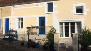 A two-story building with a yellow facade, white trim, and blue doors. There are potted plants and a small garden area in front.