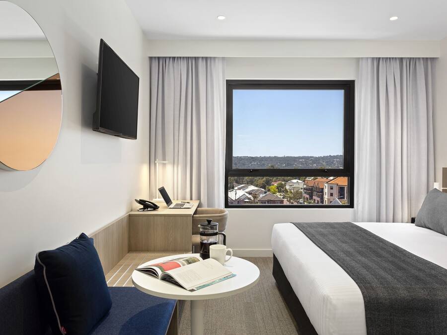 Una habitación de hotel moderna con una cama grande, un escritorio con una computadora portátil, una cafetera y vistas a la ciudad.