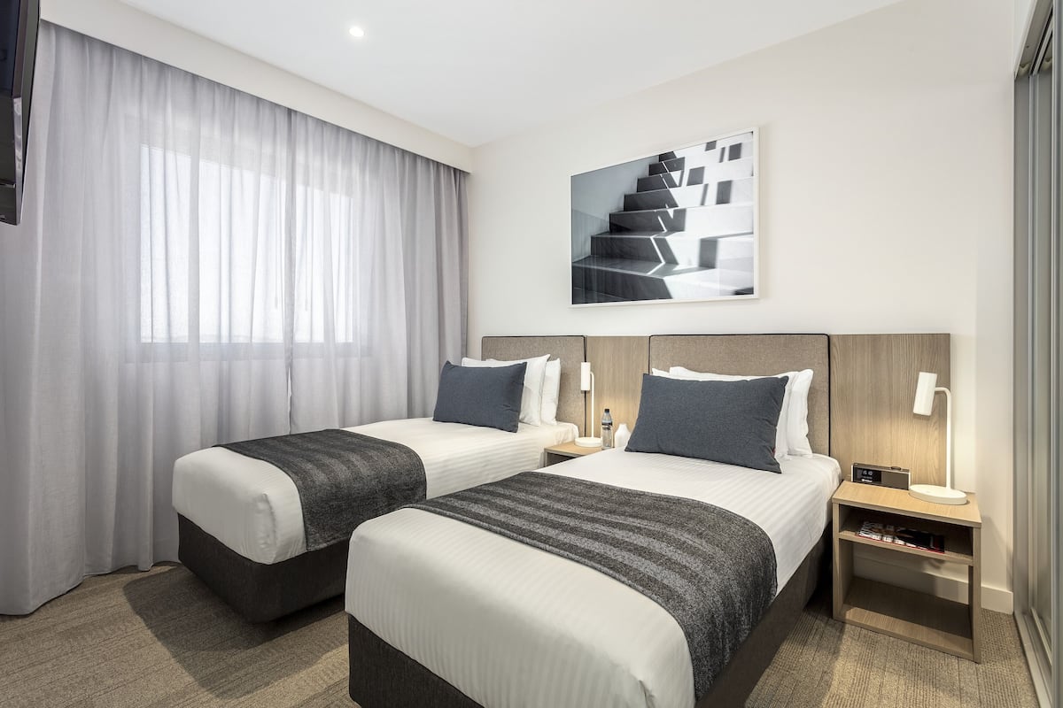 Hotellihuone, jossa on kaksi sänkyä, suuri ikkuna läpikuultavilla verhoilla, moderni taideteos seinällä sekä yöpöytä, jossa on lamppu ja pullo.