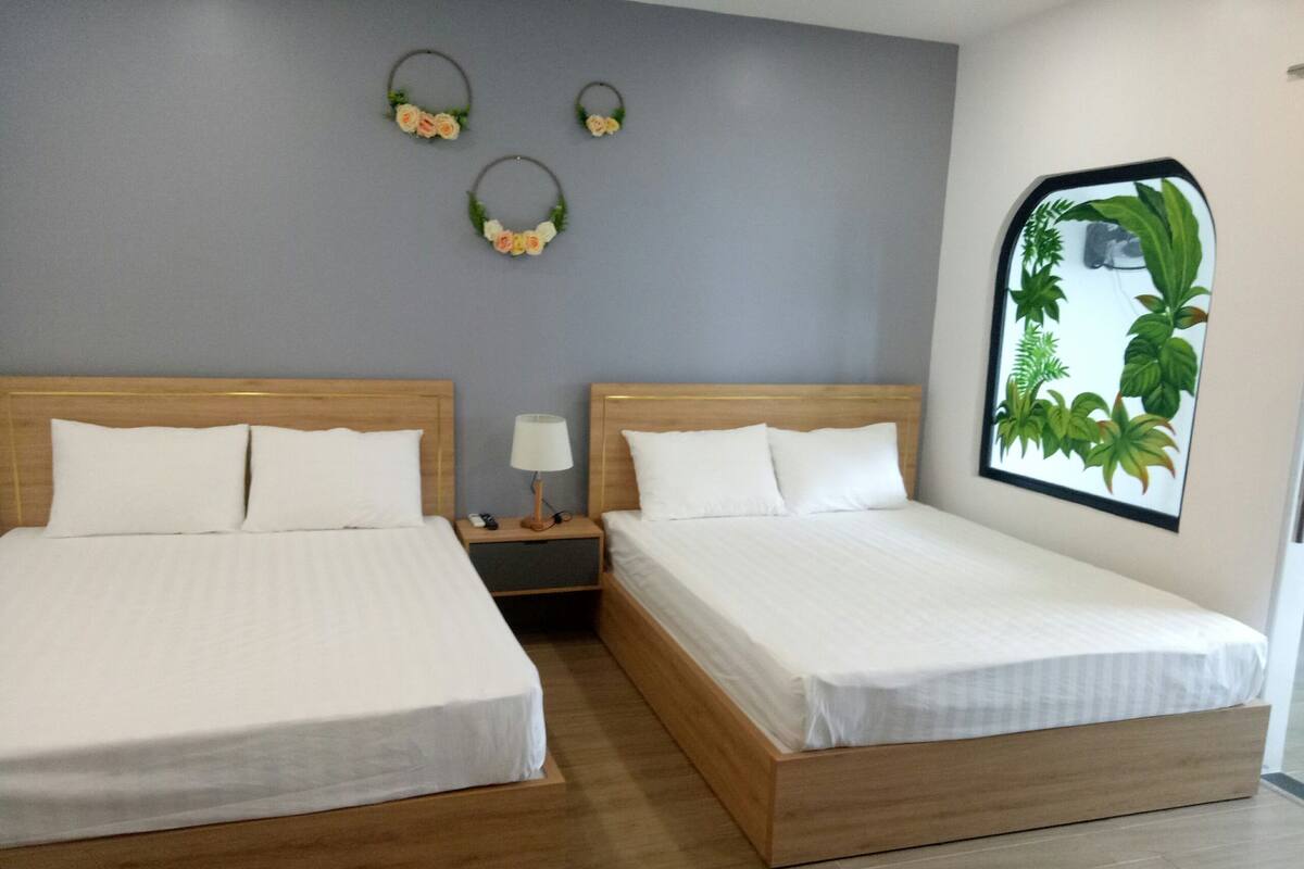 Quarto de hotel com duas camas de solteiro, mesa de cabeceira com abajur e planta fixada na parede.