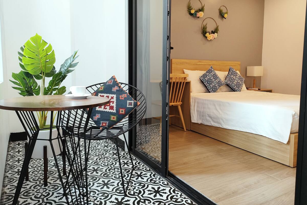 Quarto de hotel com cama, uma pequena mesa com cadeira, uma planta em vaso e piso estampado.
