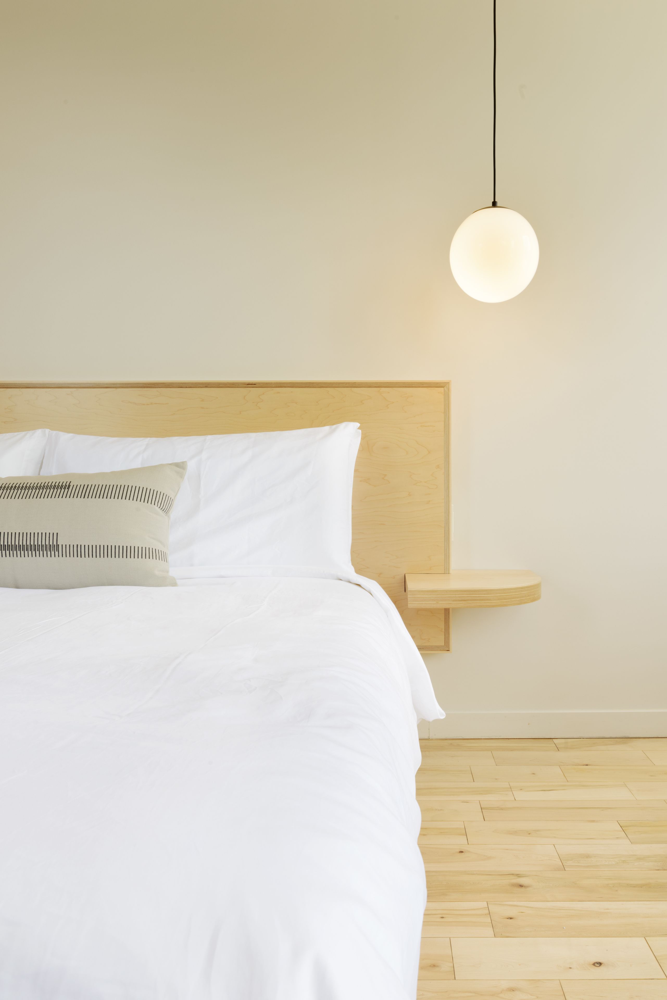 Een net opgemaakt bed met witte lakens en een enkel kussen, een houten hoofdbord en een nachtkastje met een hangende lamp.