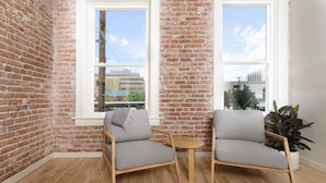 A room with exposed brick walls, two grey upholstered chairs, a small wooden table, and large windows with a view of buildings and trees. - The Jesse Hotel & Bar (Reno)