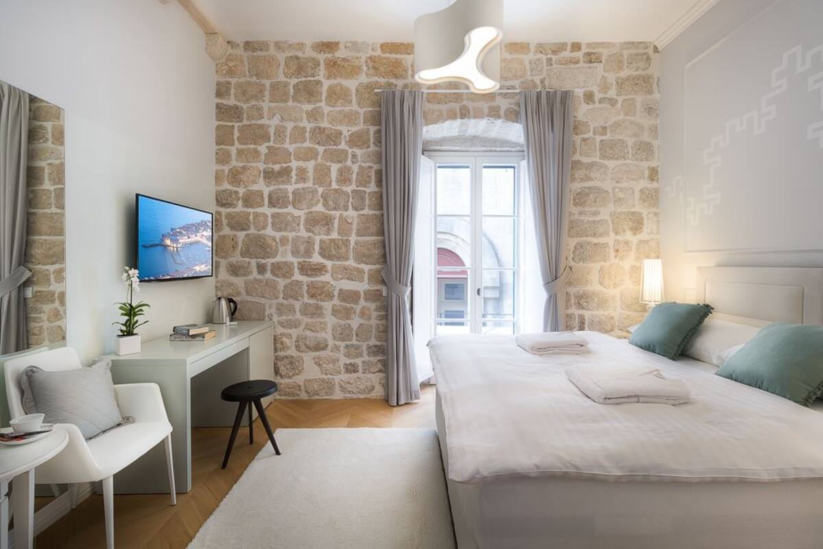 A modern bedroom with a stone wall, a bed with white linens, a desk with a chair, and a television on the wall.