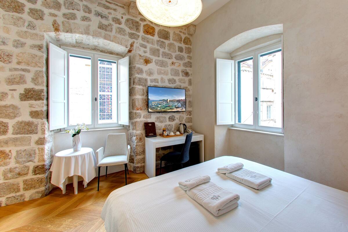 A room with stone walls, a bed with towels, a desk with a computer, and a TV on the wall.