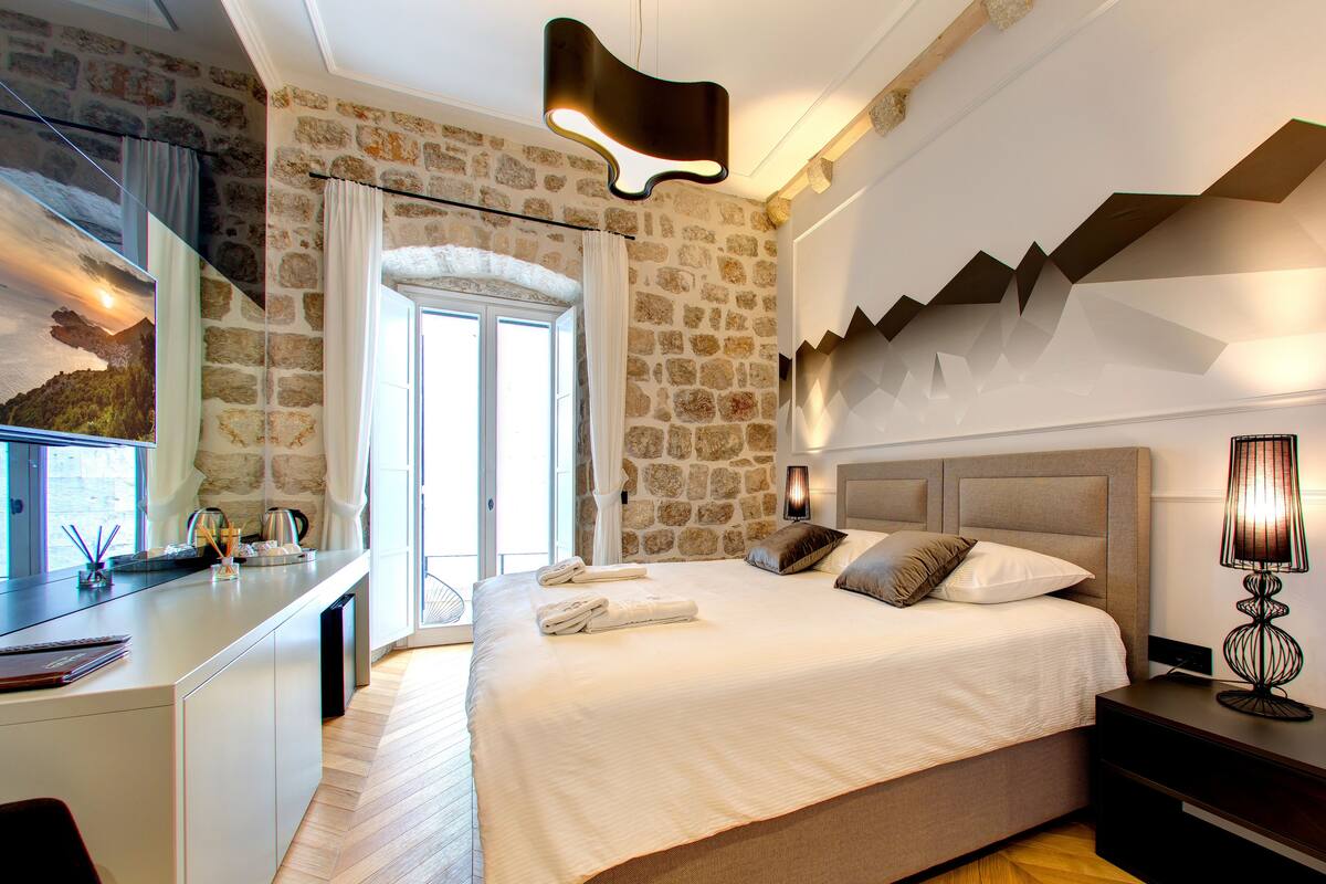 A bedroom with a stone wall, a bed with pillows, a desk with a mirror, and a lamp.