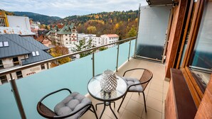 Varanda com mesa de vidro e duas cadeiras, com vista para uma paisagem urbana com árvores e edifícios em tons de outono.