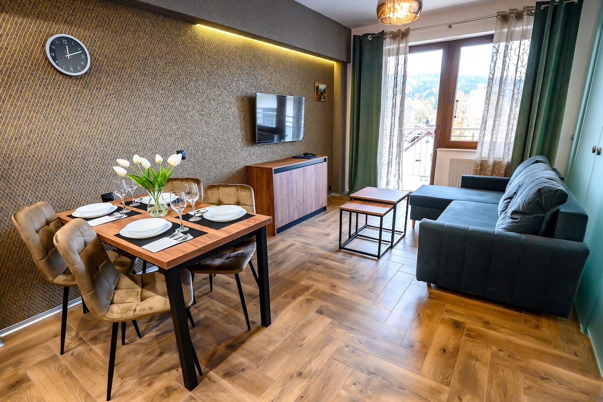 Un salon moderne avec une table à manger en bois, préparée pour quatre personnes, un canapé et un téléviseur fixé au mur.