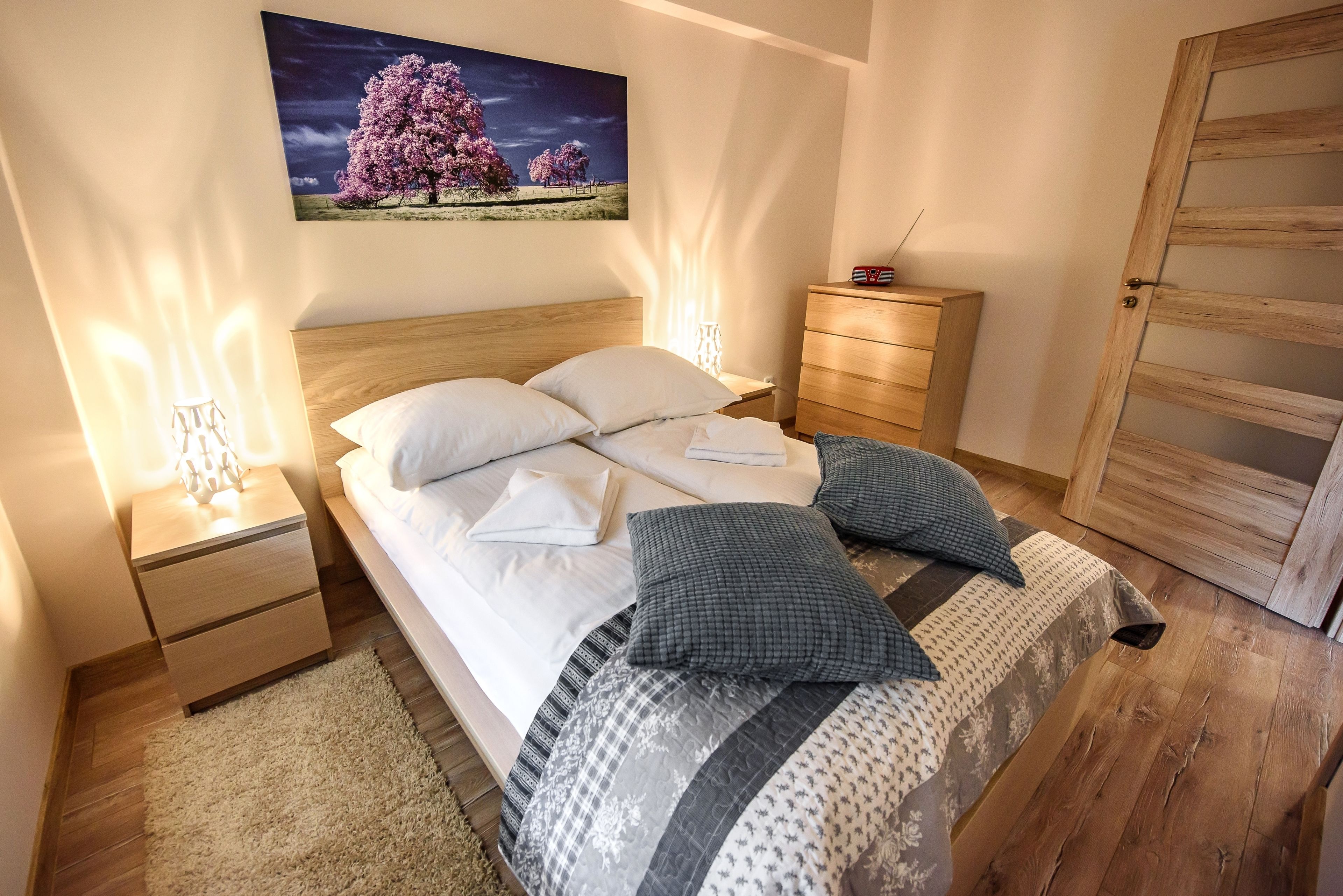 A neatly made bed with pillows and a blanket, a bedside table with a lamp, a wooden nightstand, a dresser, and a painting on the wall.