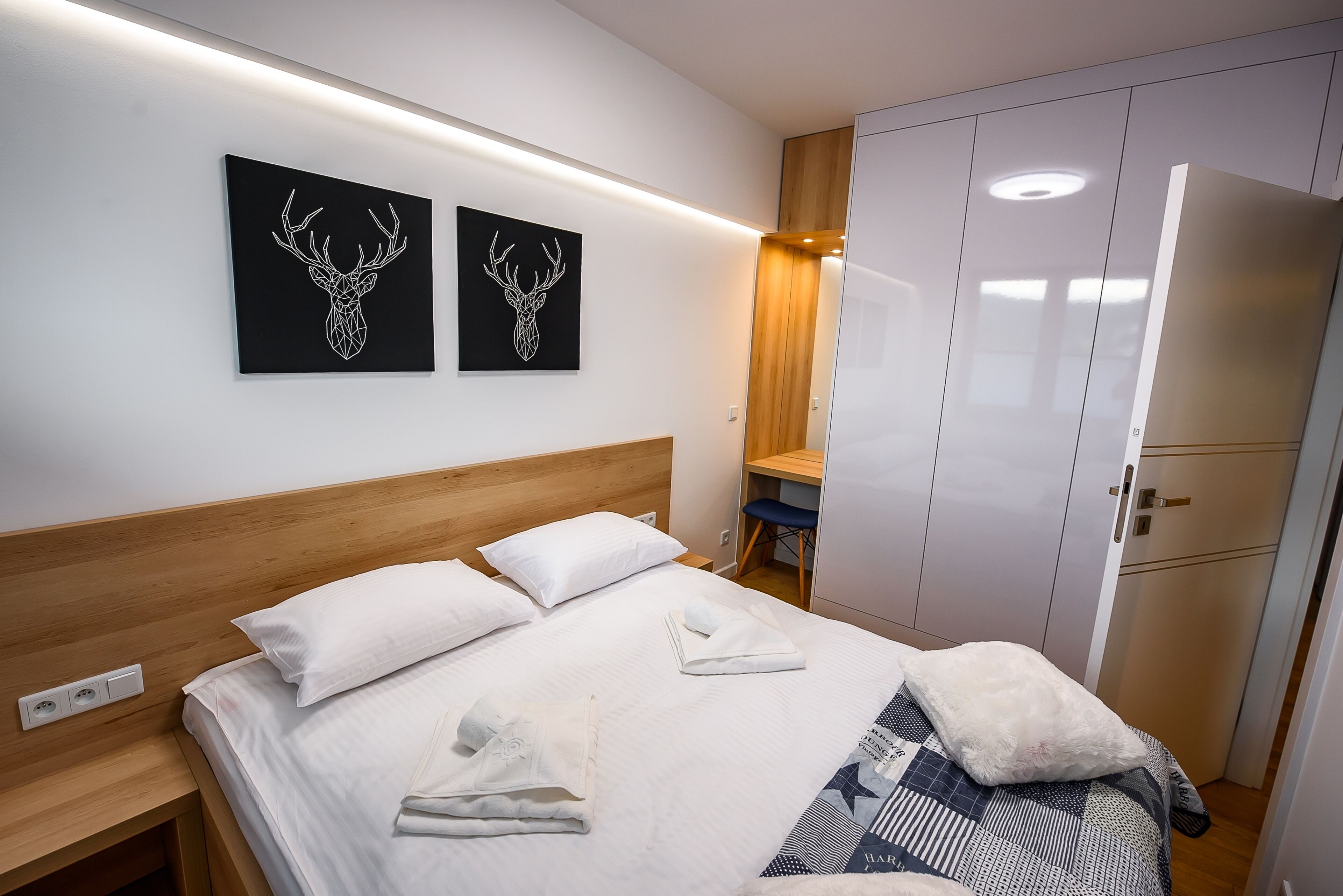 A neatly made bed with white linens and a folded towel, a wooden headboard, and two wall-mounted artwork of deer antlers.