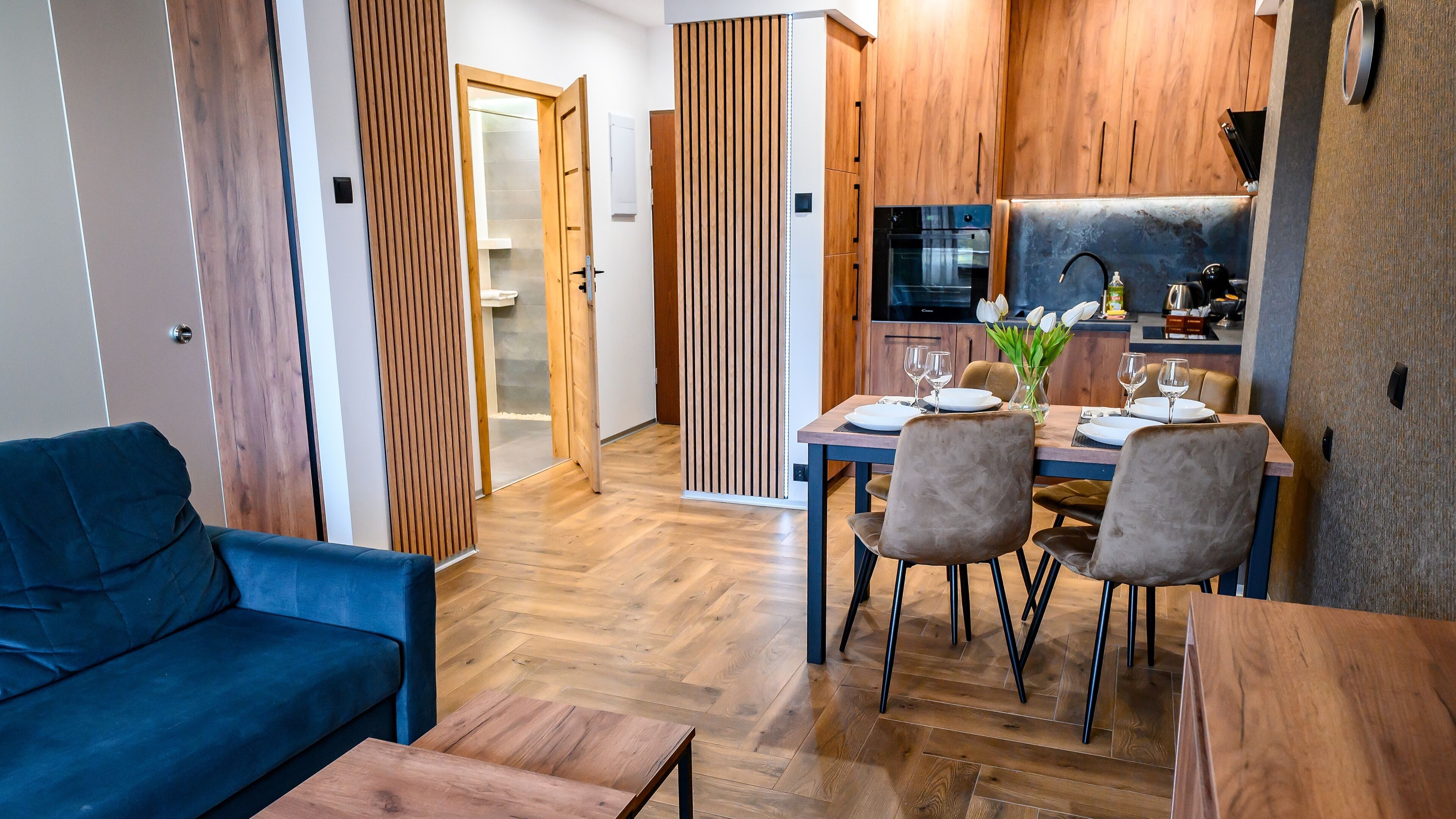 A modern kitchen with wooden cabinets, a dining table set for four, and a blue sofa in the living area.