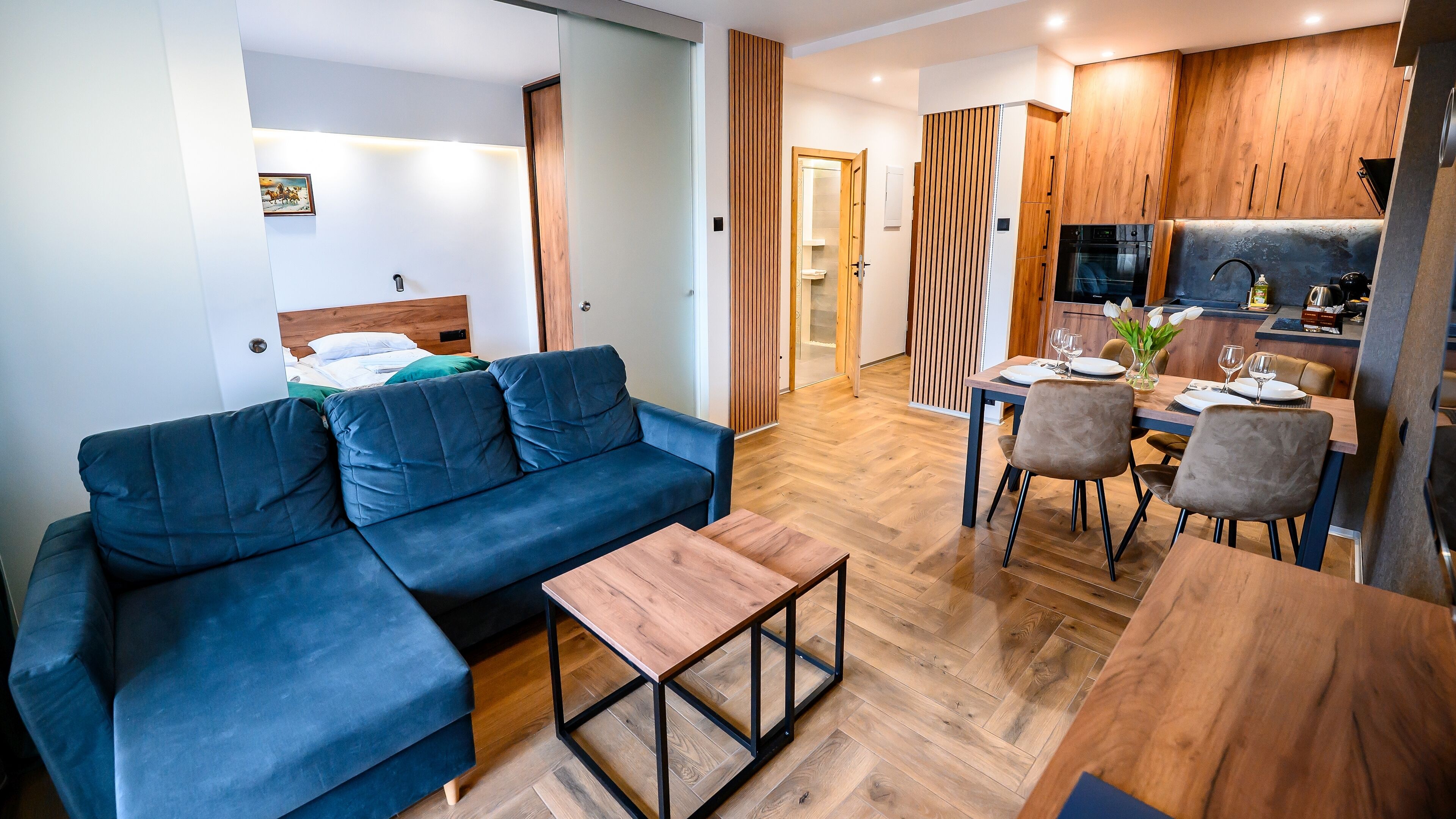 A modern living room with a blue sofa, a wooden coffee table, and a dining area with a wooden table and chairs.