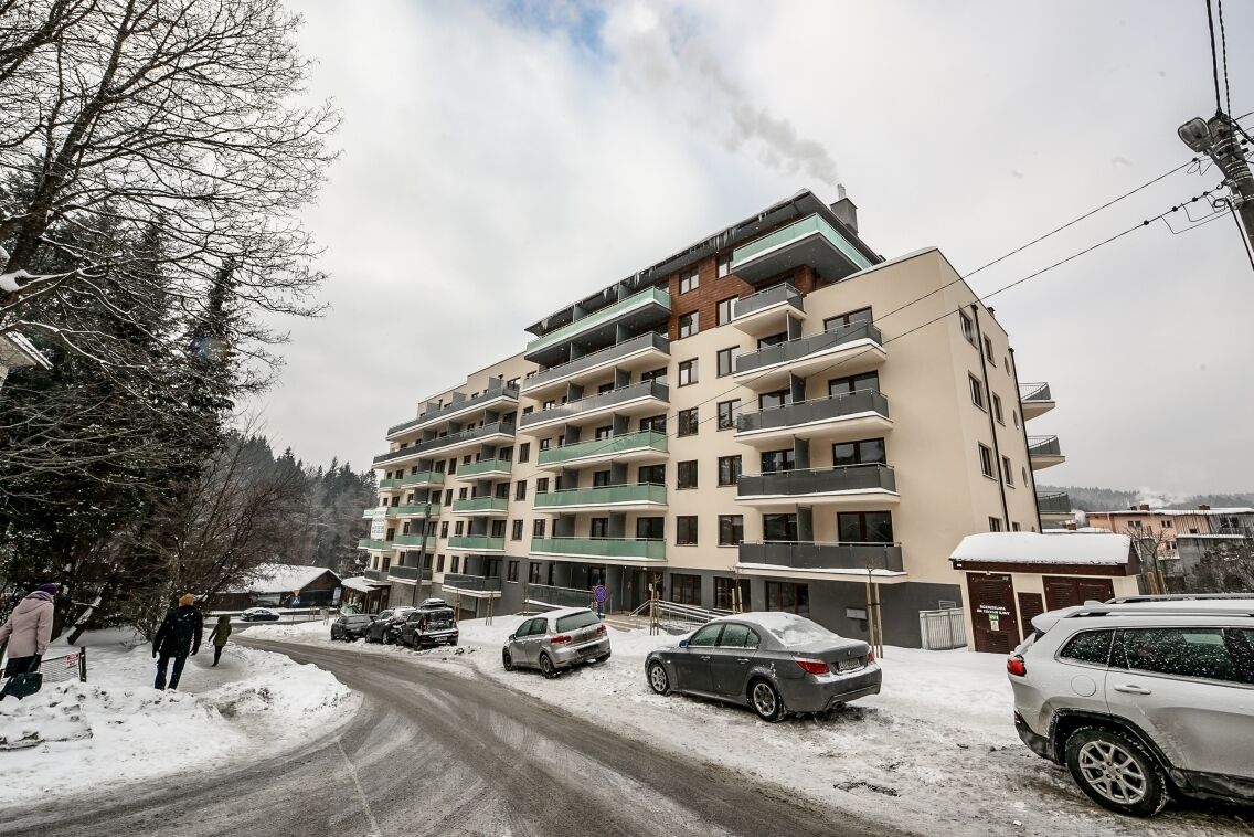 A multi-story residential building with balconies, a snow-covered street, and parked cars.