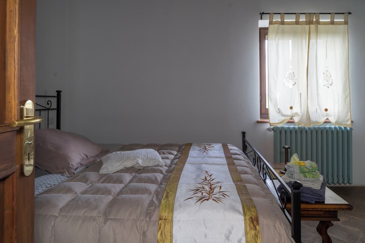 Et soveværelse med seng, et vindue med gardiner, en radiator og et natbord med genstande.