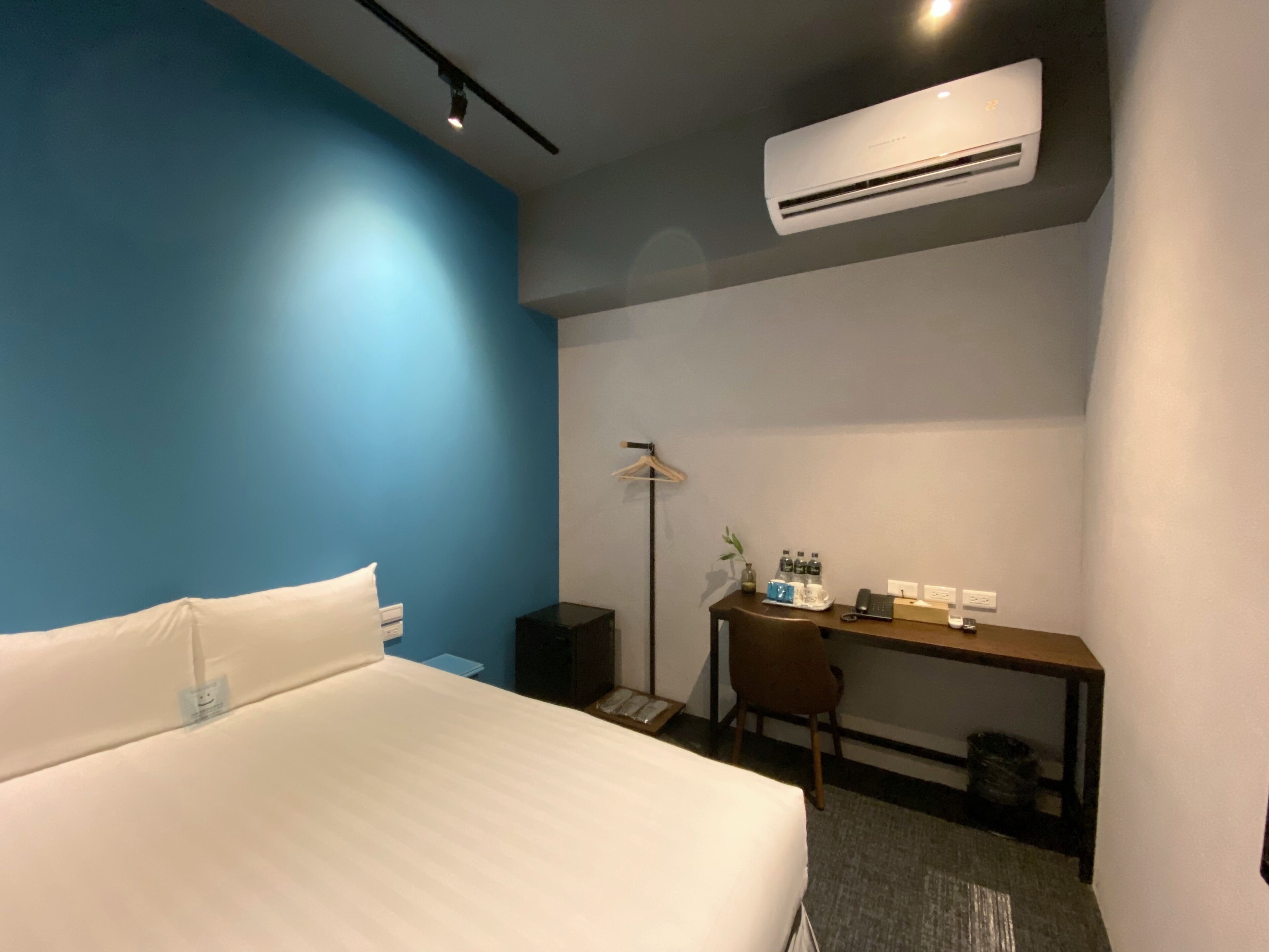 A hotel room with a bed, a desk, a chair, an air conditioner, a lamp, and a small plant.