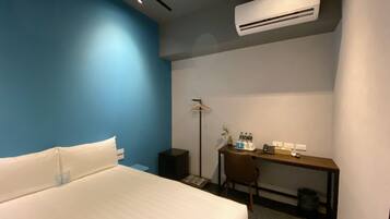 A hotel room with a bed, a desk, a chair, an air conditioner, a lamp, and a small plant.