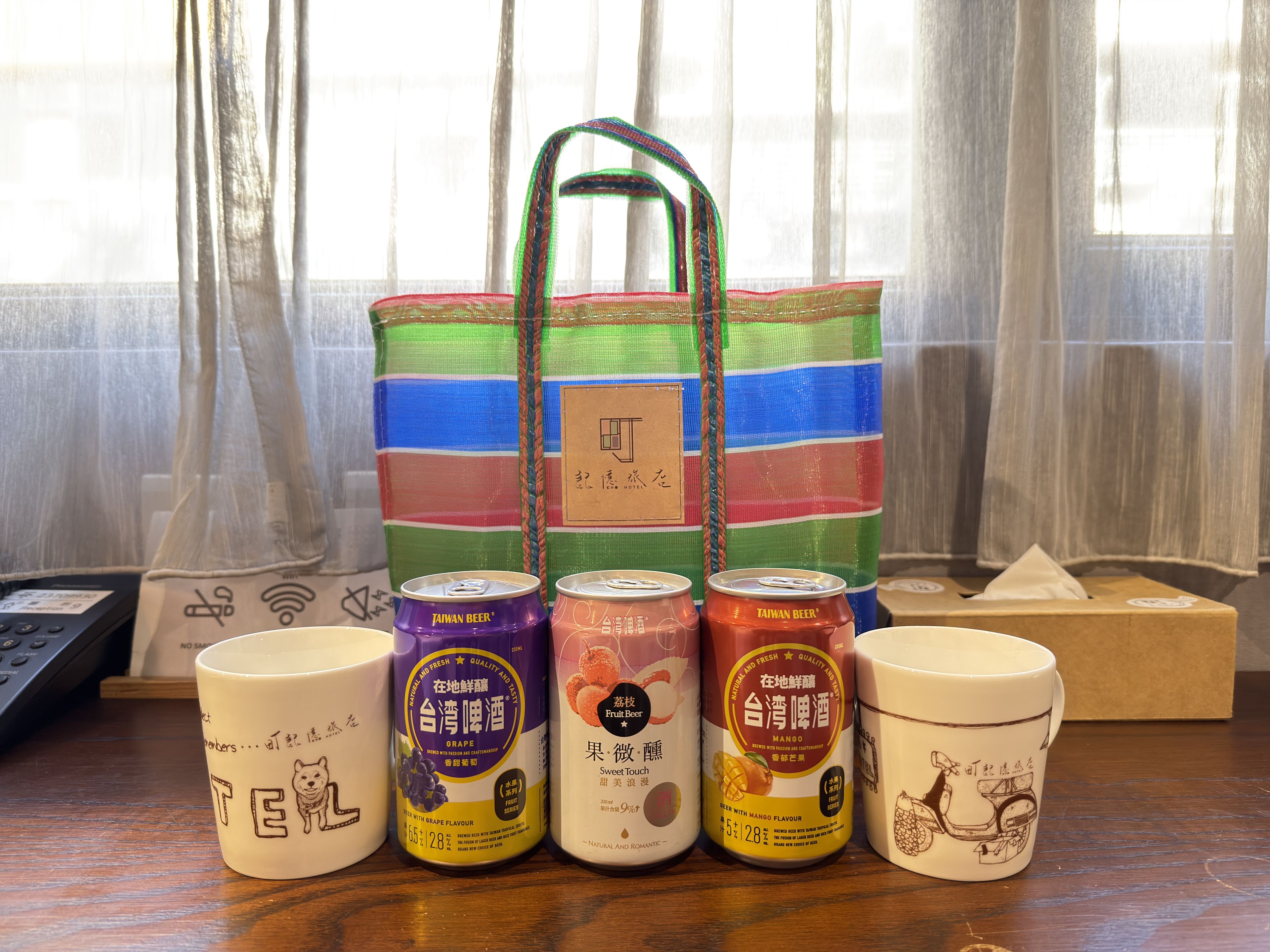 A table with a colorful tote bag, three cans of Taiwan beer, and two mugs with illustrations.