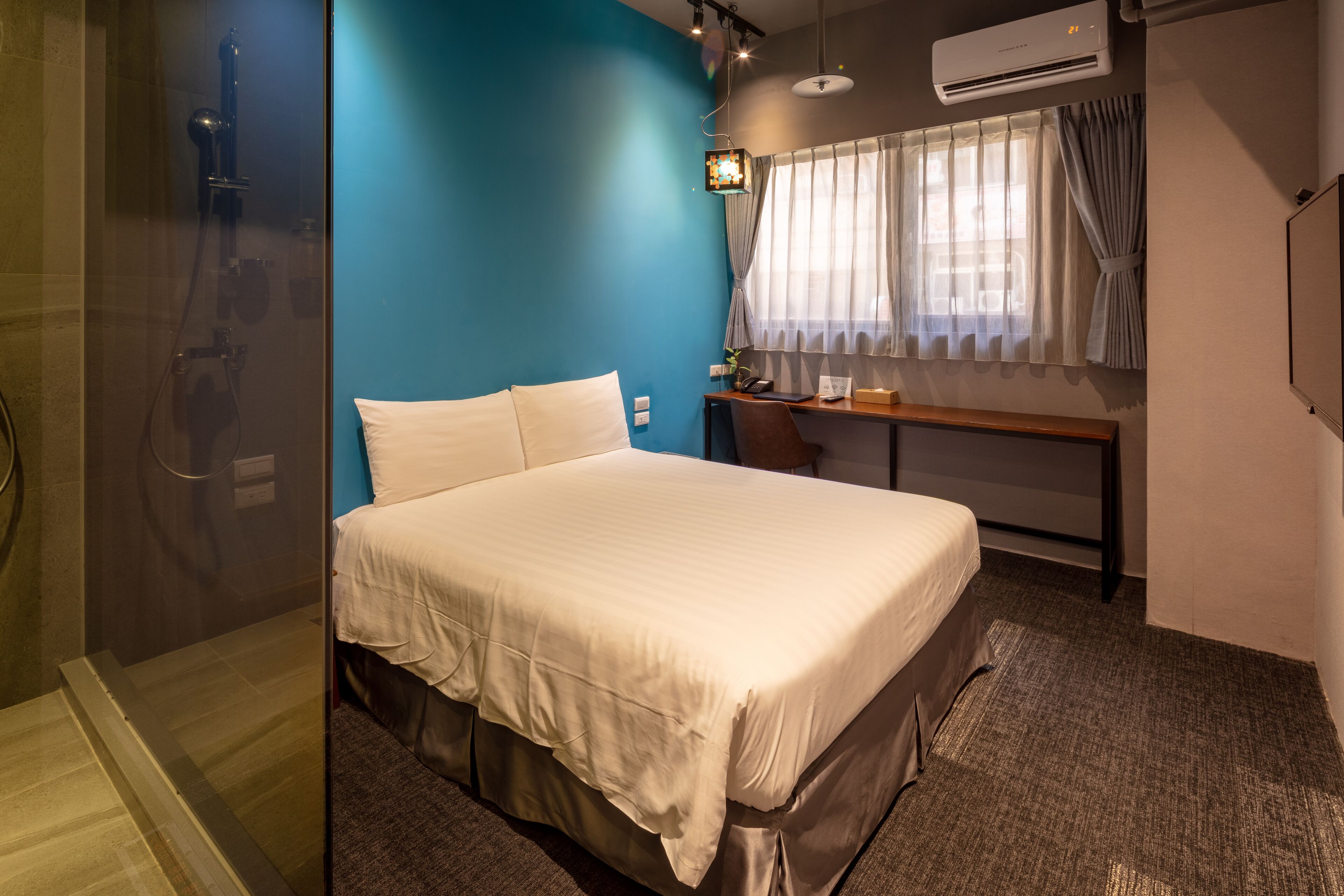 A hotel room with a bed, a desk, a chair, a shower, and an air conditioning unit.