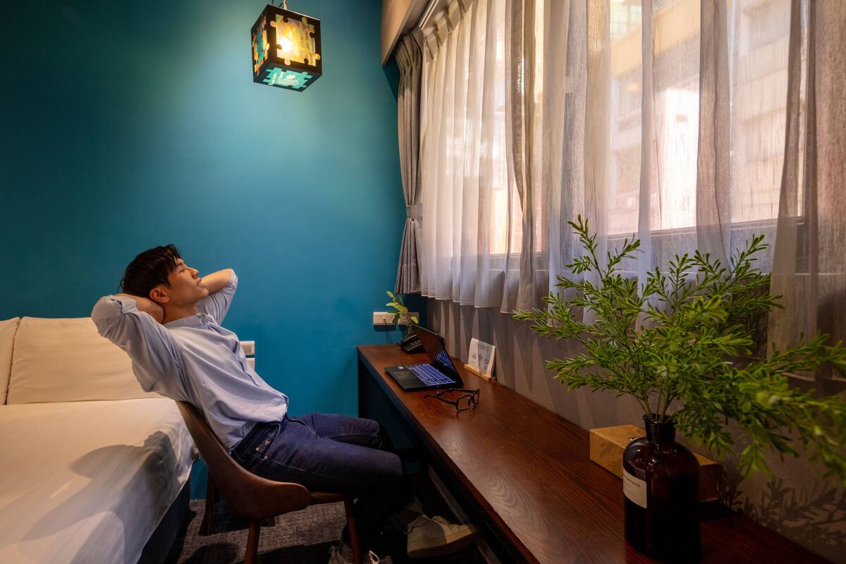 A room with a bed, a desk with a laptop, a potted plant, and a window with sheer curtains.