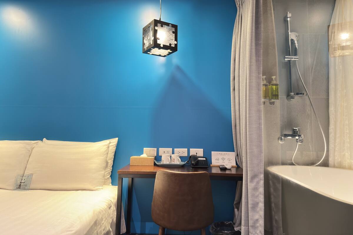 A room with a bed, a desk with a chair, a shower area, and a hanging light fixture.