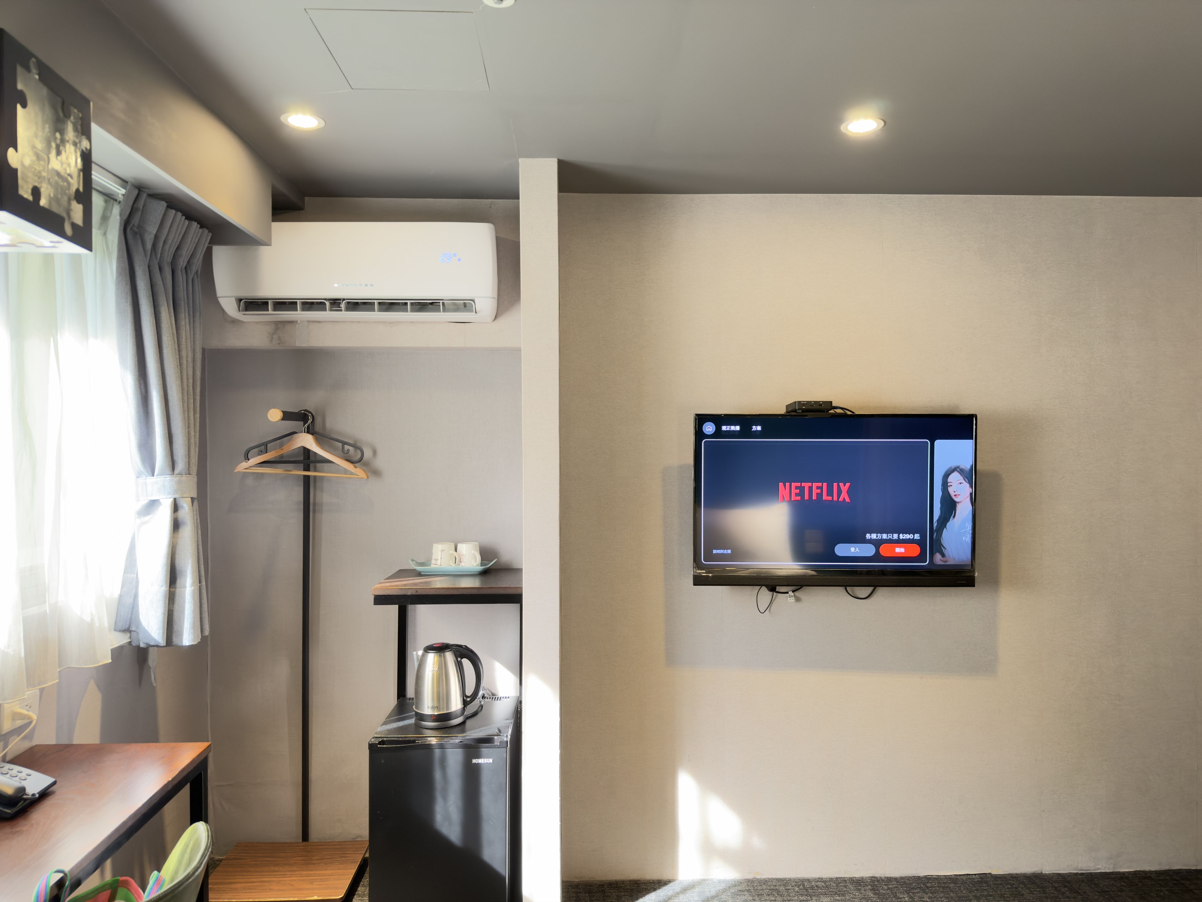 A hotel room with a flat-screen TV displaying the Netflix logo, a small kitchenette with a coffee maker, a wooden table, and a wall-mounted TV.
