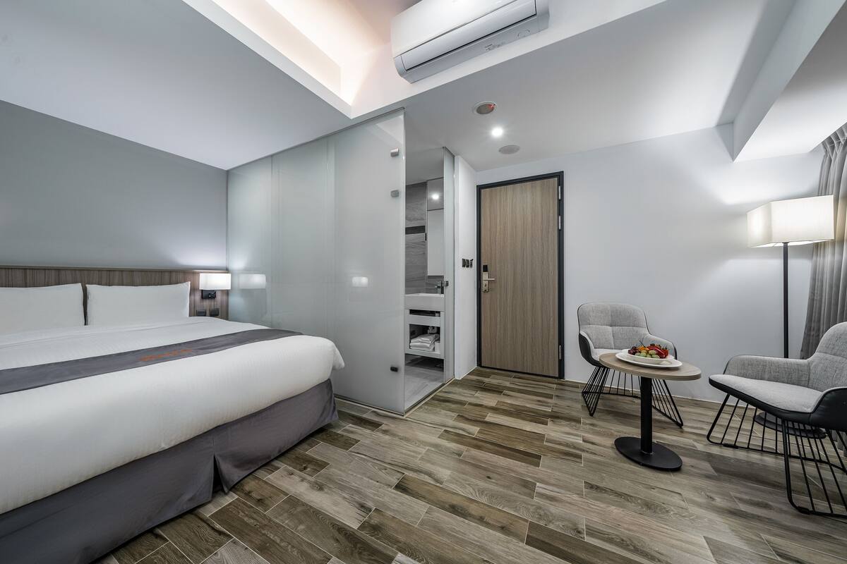 Ein modernes Hotelzimmer mit einem großen Bett, einem kleinen Esstisch, zwei Stühlen, einem Kleiderschrank und einer Deckenleuchte.