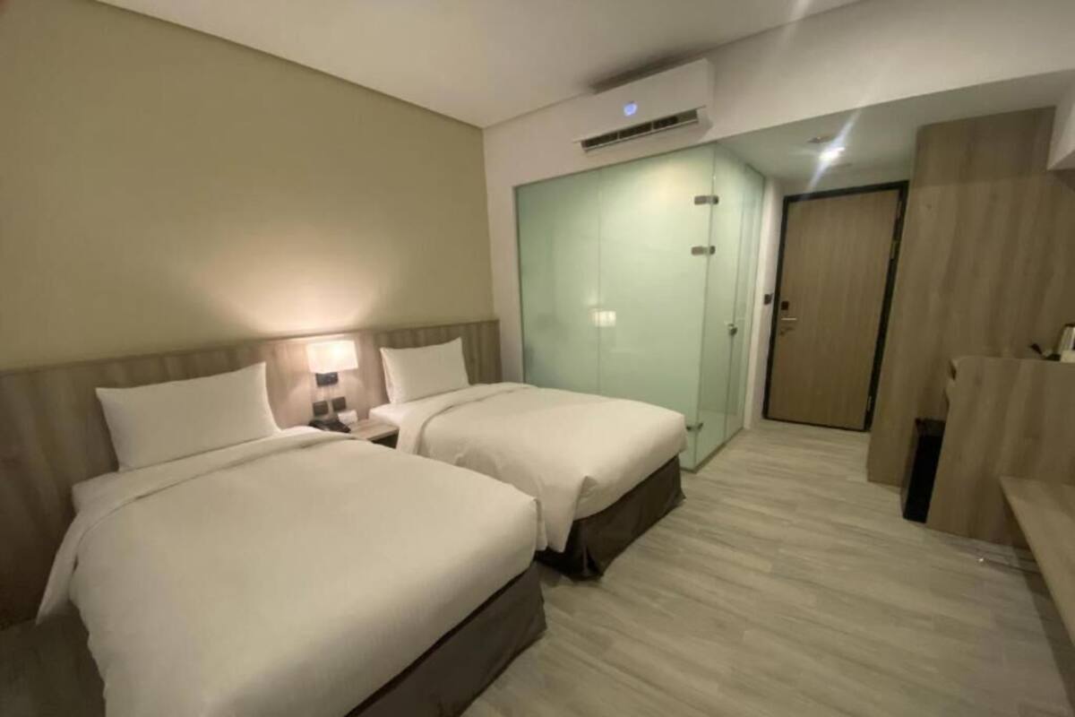 Ein Hotelzimmer mit zwei Betten, einer Glastrennwand, einem Kleiderschrank und einem Badezimmer.